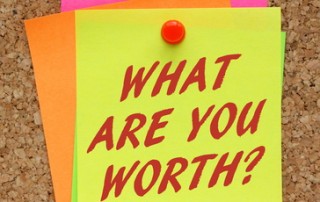 The question What Are You Worth in red text on a yellow sticky note as a reminder to assess your value in terms of financial rewards
